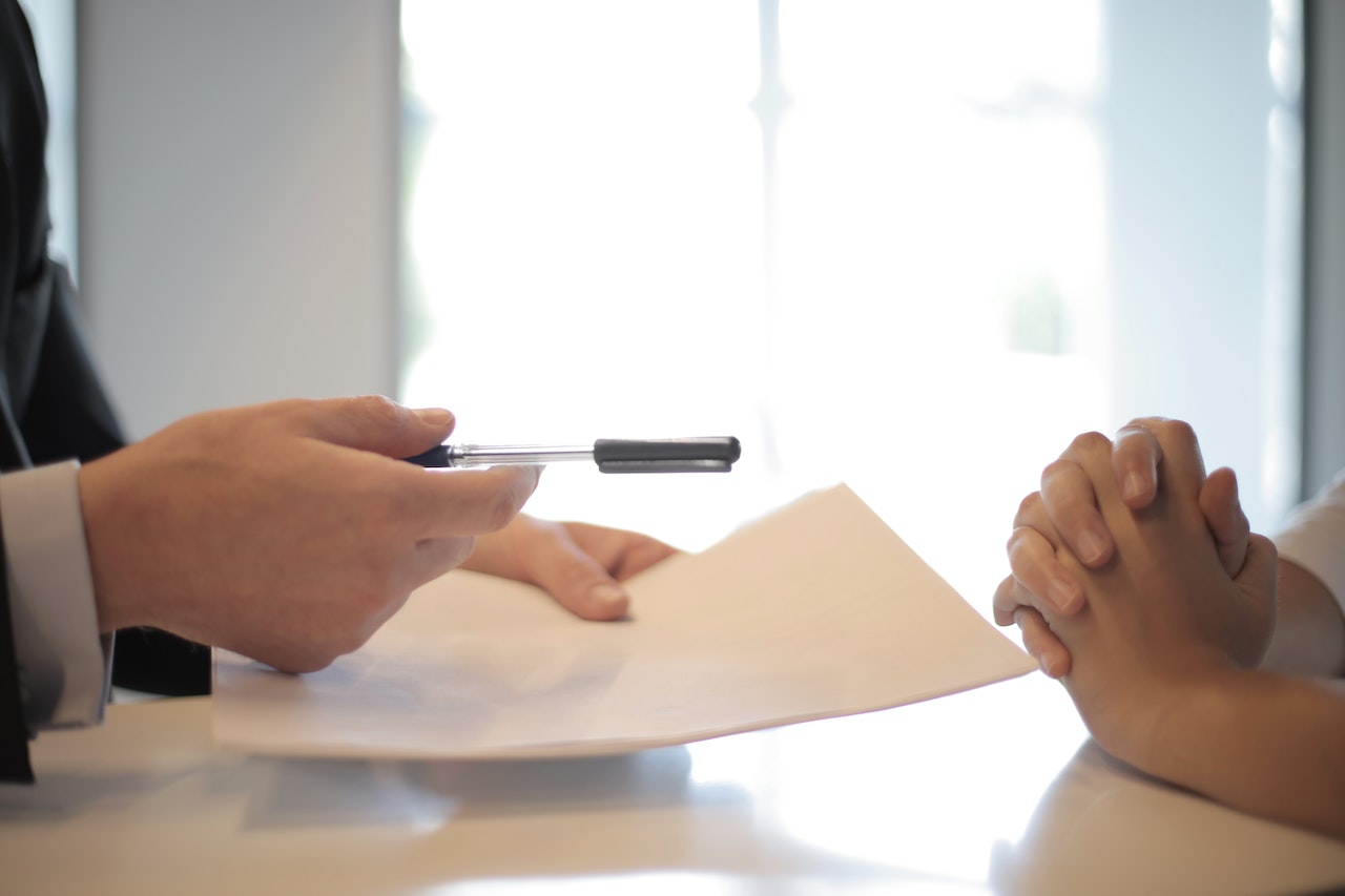 A photo of a man holding a paper sheet and offering a pen to another person, symbolizing the signing of documents related to legal compliance for remote workers